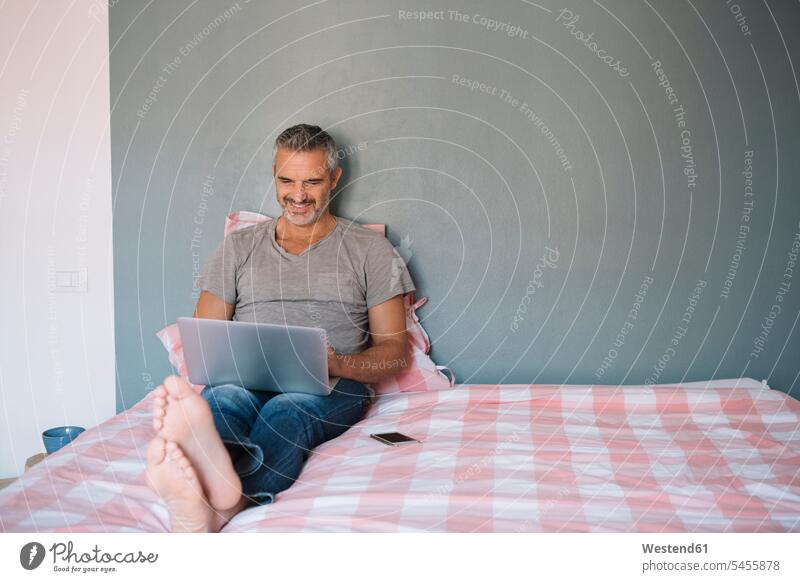Smiling mature man sitting on bed at home using laptop Laptop Computers laptops notebook men males use smiling smile Seated beds computer computers Adults