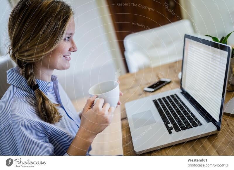 Smiling woman sitting at desk with cup of coffee and laptop desks Seated smiling smile Coffee Laptop Computers laptops notebook females women Table Tables Drink