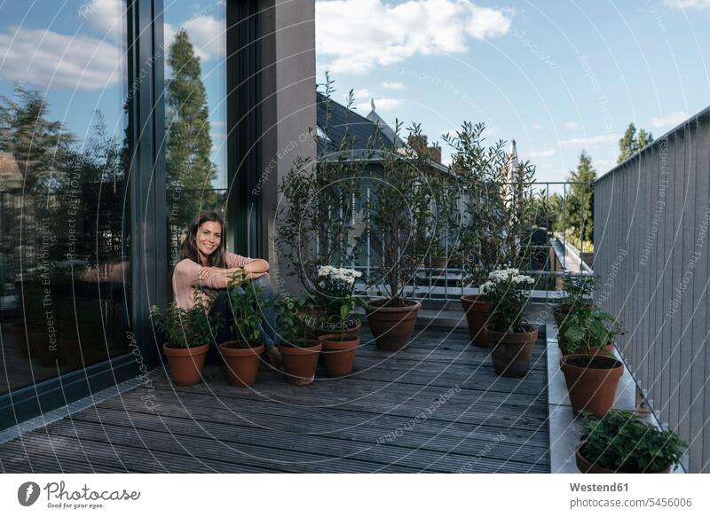 Smiling woman relaxing on balcony surrounded by plants relaxed relaxation Plant Plants sitting Seated balconies smiling smile females women Adults grown-ups