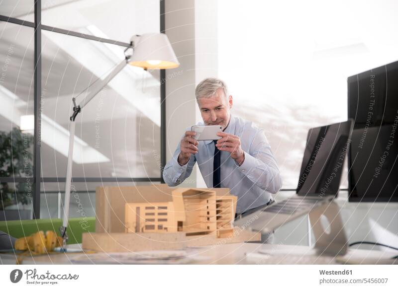 Businessman taking pictures of architectural model sitting Seated planning office Smartphone iPhone Smartphones working At Work expertise competence competent