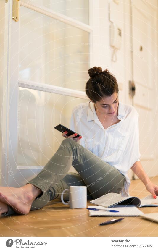 Young woman sitting on the floor with coffee mug and smartphone looking at booklet females women Adults grown-ups grownups adult people persons human being
