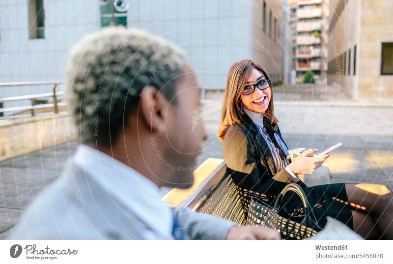 Young businessman and woman sitting on bench, talking business people businesspeople on the move on the way on the go on the road speaking benches flirting