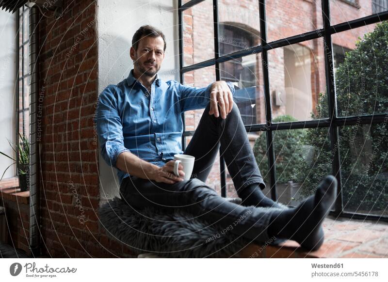 Mature man sitting on window sill, relaxing with cup of coffee home at home Coffee Cup Coffee Cups windowsill sills window sills Window Cill windowsills Seated