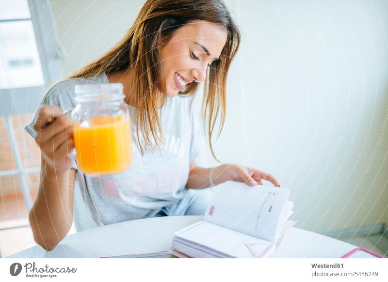 Smiling young woman with glass of orange juice looking at agenda females women home office working from home home business schedule timetable Adults grown-ups