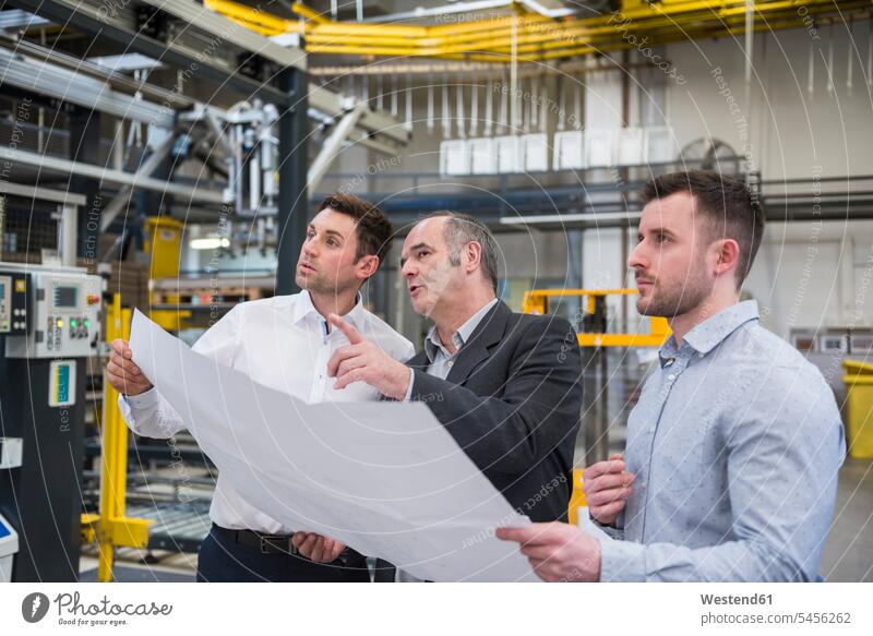 Three men with blueprint talking in factory shop floor factories man males speaking colleagues working At Work Adults grown-ups grownups adult people persons