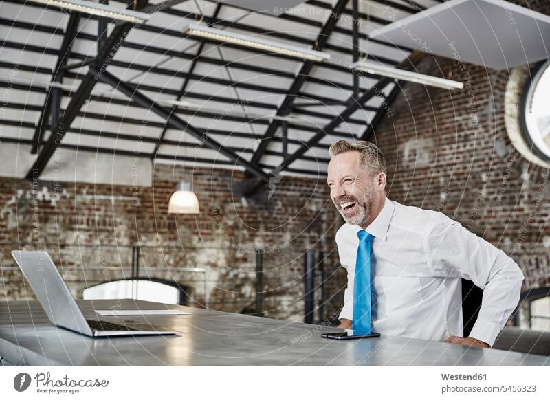 Laughing businessman with laptop sitting at table in a loft Seated Laptop Computers laptops notebook laughing Laughter Businessman Business man Businessmen