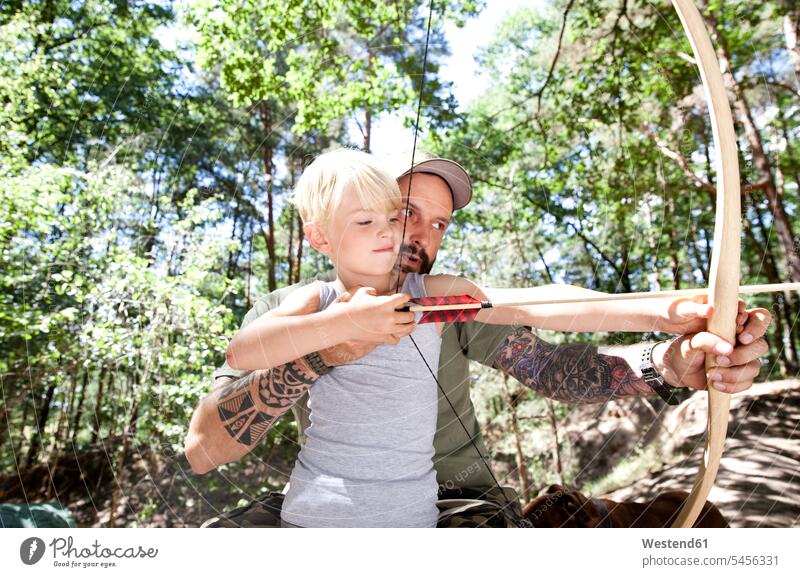 Father and son shooting with bow and arrow in the forest sons manchild manchildren woods forests aiming father pa fathers daddy dads papa arrows boy boys males