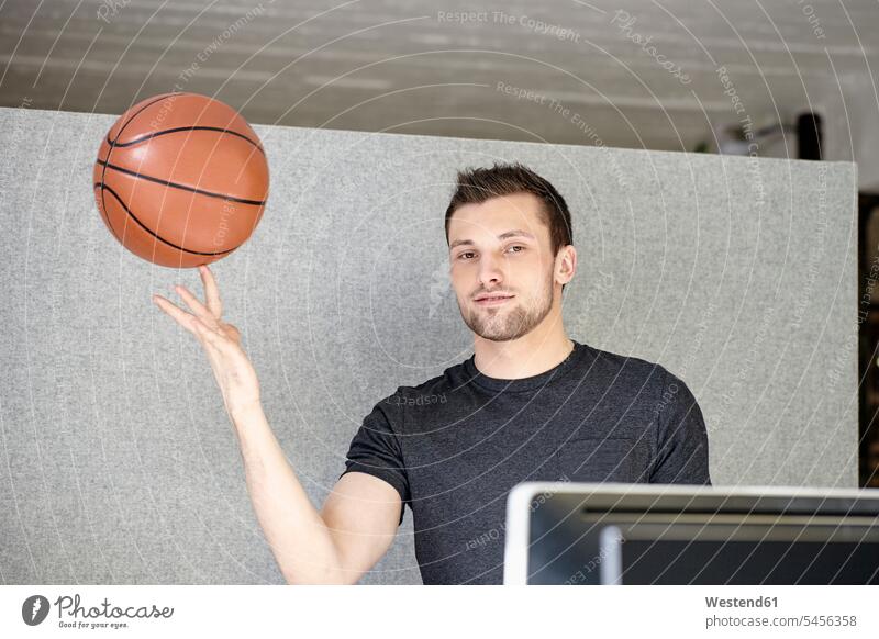Young man working in office, balancing a basketball smiling smile young man young men balls sitting Seated Basketball balance offices office room office rooms
