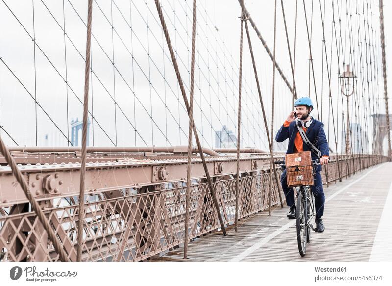 USA, New York City, man on bicycle on Brooklyn Bridge using cell phone New York State mobile phone mobiles mobile phones Cellphone cell phones men males