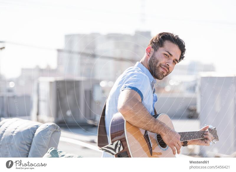 Young man on rooftop sitting on sofa and playing guitar Seated musician musicians couch settee sofas couches settees guitars roof terrace deck cheerful gaiety