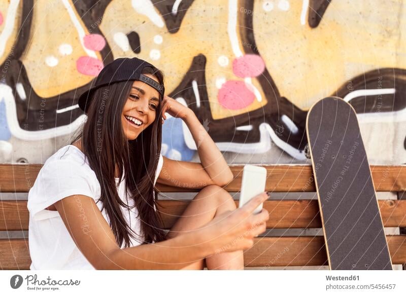 Smiling young woman with skateboard looking at cell phone mobile phone mobiles mobile phones Cellphone cell phones females women bench benches laughing Laughter