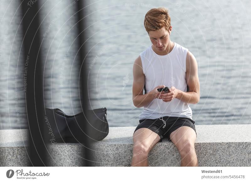 Young athlete sitting on a wall at riverbank with cell phone and earphones exercising exercise training practising Jogging walls bag bags mobile phone mobiles