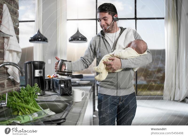 Father with headset making coffee in kitchen holding baby father pa fathers daddy dads papa Coffee domestic kitchen kitchens parents family families people