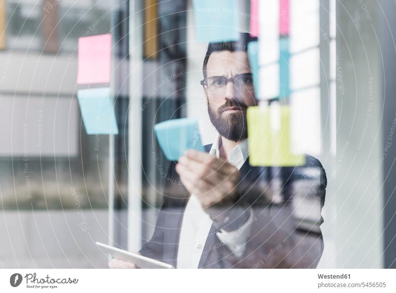 Young businessman attaching adhesive notes to glass pane glass panes gluing glueing glued serious earnest Seriousness austere Adhesive Note sticky note