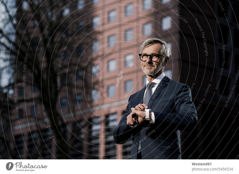 Grey-haired businessman in front of red skyscraper pointing on his smartwatch Businessman Business man Businessmen Business men smart watch skyscrapers