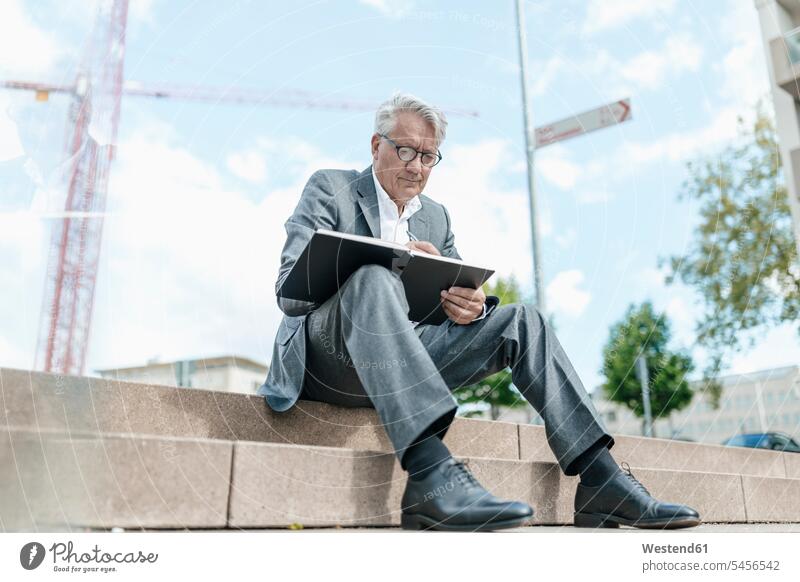 Portrait of senior businessman sitting on stairs with notebook in front of construction crane Businessman Business man Businessmen Business men business people