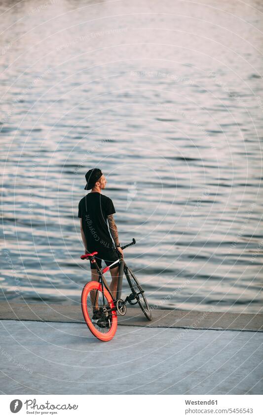 Young man with fixie bike at the waterfront River Rivers standing bicycle bikes bicycles men males waters body of water Adults grown-ups grownups adult people