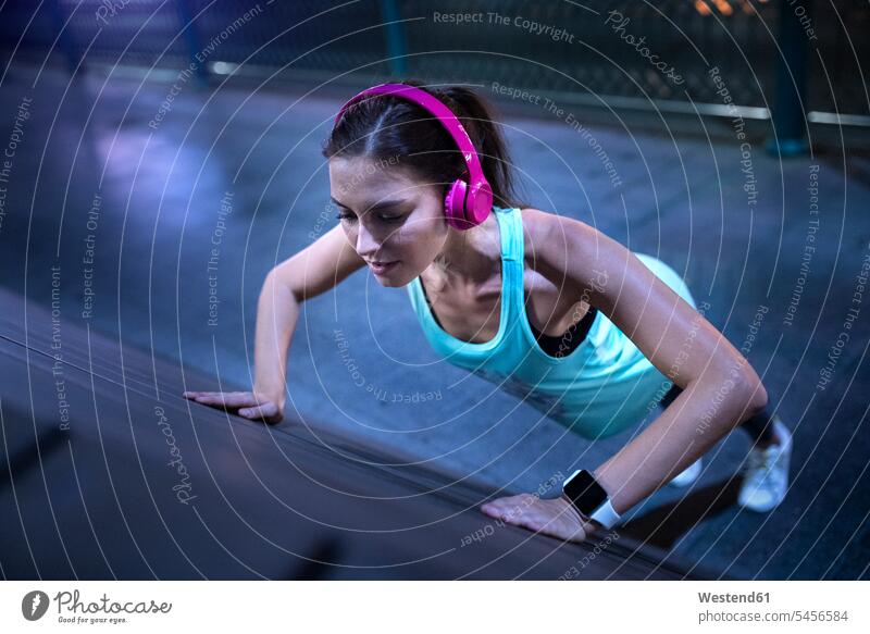 Young woman with pink headphones doing pushups in modern urban setting at night exercising exercise training practising Listening Music headset fit urbanity
