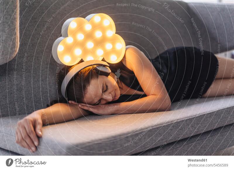 Woman lying on couch wearing headphones with illuminated cloud above her laying down lie lying down headset relaxed relaxation woman females women relaxing