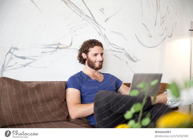 Smiling young man sitting on couch in a coffee shop using laptop cafe Laptop Computers laptops notebook men males computer computers Adults grown-ups grownups