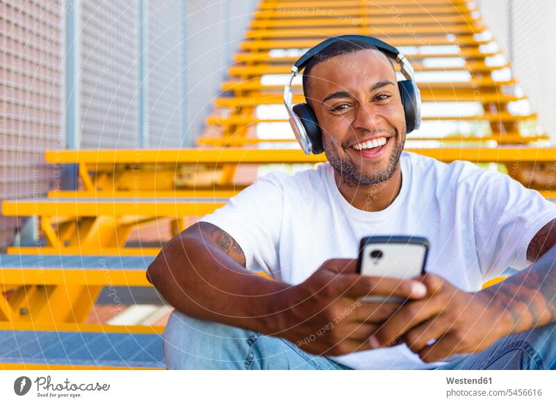 Portrait of laughing young man with headphones and smartphone sitting on stairs Smartphone iPhone Smartphones portrait portraits men males headset mobile phone