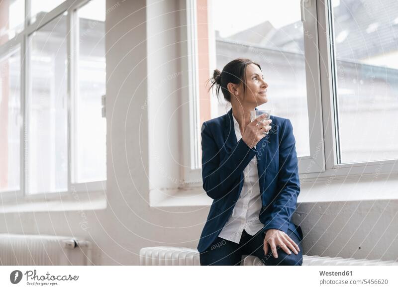 Businesswoman with glass of water sitting on radiator in a loft looking through window businesswoman businesswomen business woman business women females