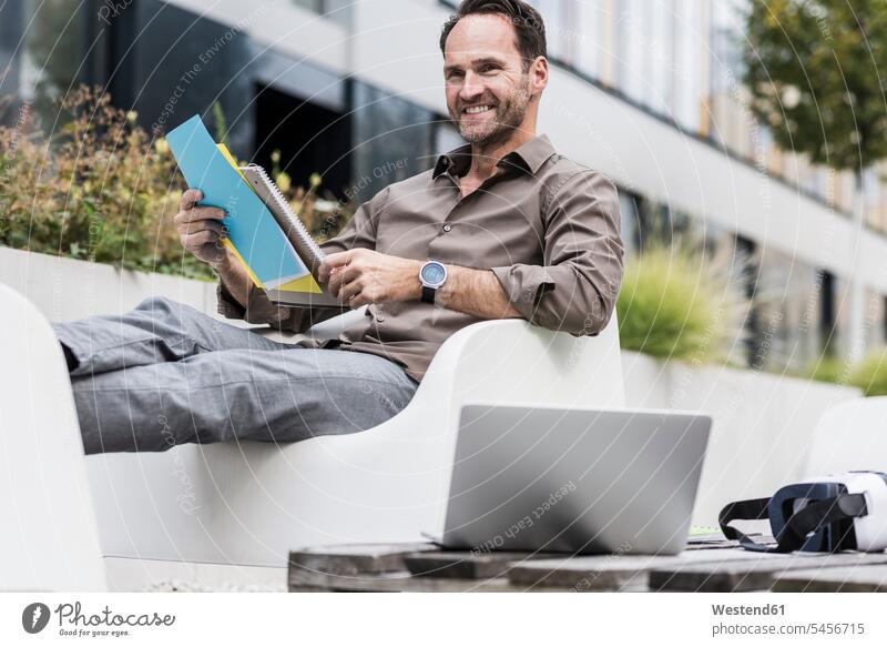 Smiling businessman with document, laptop and Virtual Reality Glasses sitting on terrace Businessman Business man Businessmen Business men business people