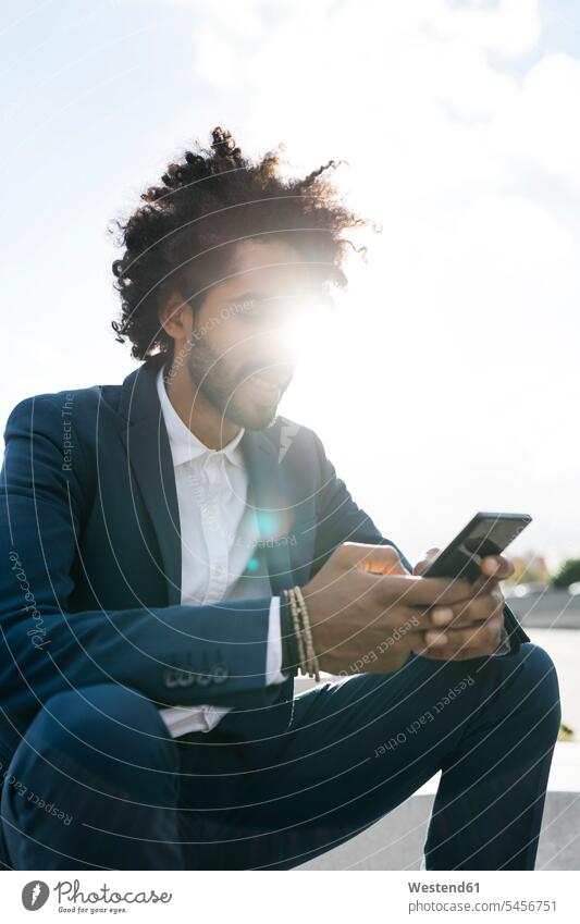 Young businessman sitting outdoors in sunshine using cell phone Businessman Business man Businessmen Business men males Sunny Day sunny Seated mobile phone