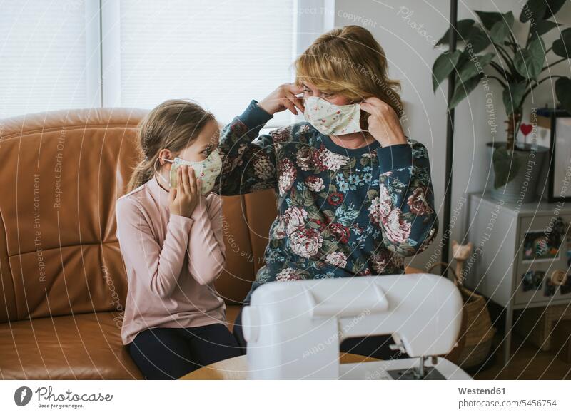 Grandmother and granddaughter wearing protective face masks while sitting on sofa in living room at home color image colour image Germany indoors indoor shot