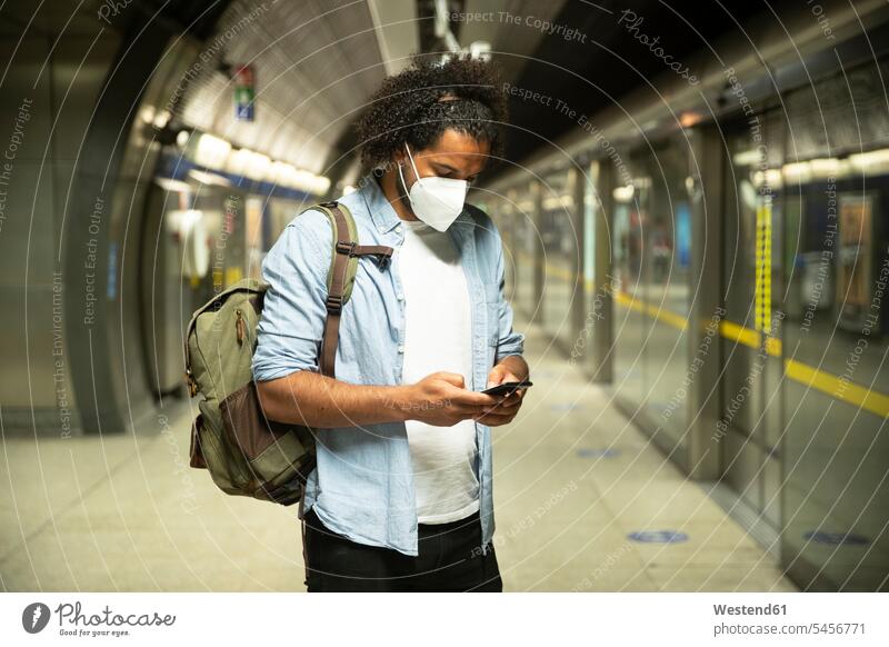 Young man wearing protective mask standing at underground station platform, London, UK back-pack back-packs backpacks rucksack rucksacks transport railroad