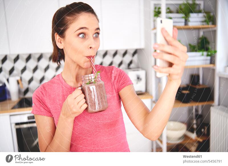 Portrait of young woman taking selfie with smartphone in the kitchen while drinking smoothie domestic kitchen kitchens portrait portraits females women Selfie