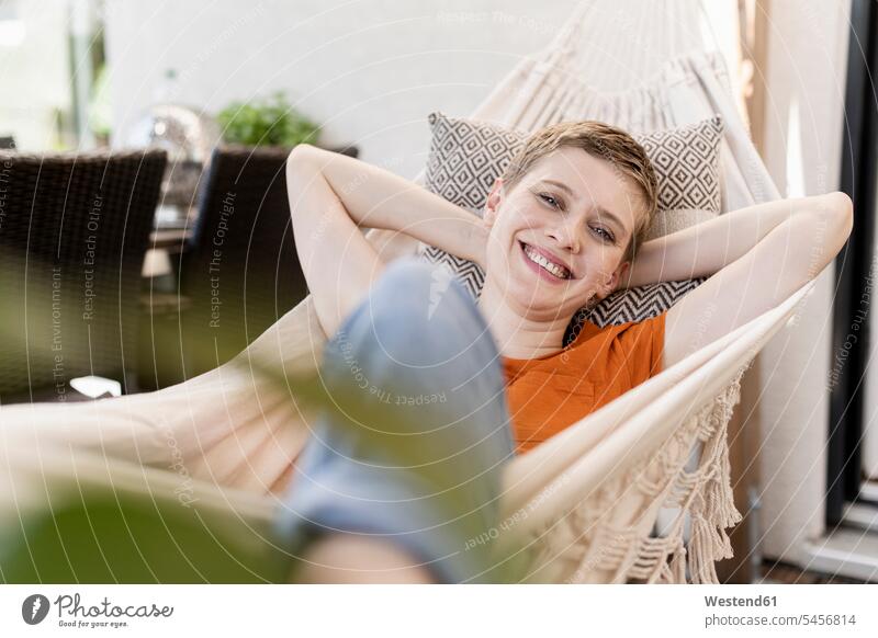 Smiling mid adult woman with hands behind head resting on hammock in porch color image colour image Germany leisure activity leisure activities free time