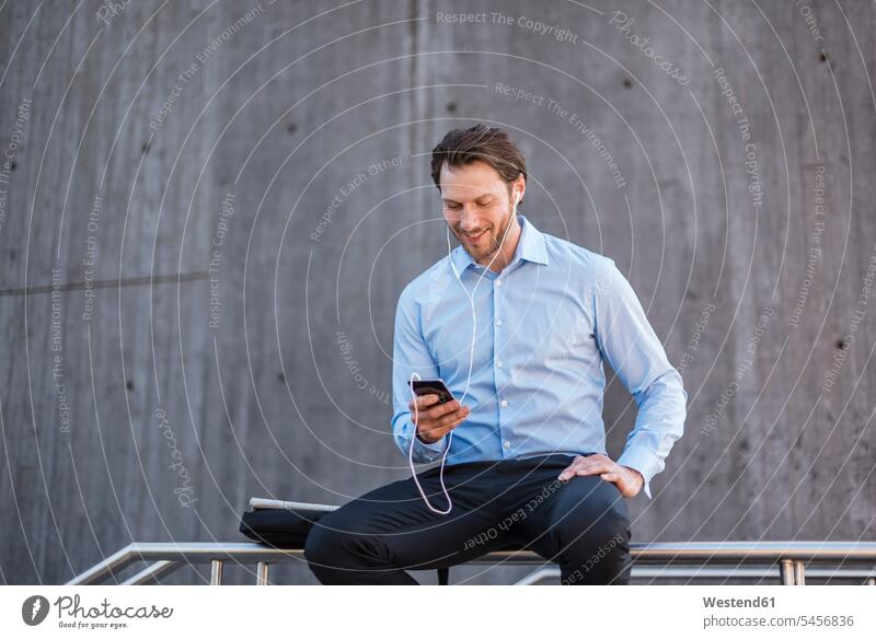 Smiling businessman with earbuds sitting on a railing looking at smartphone Businessman Business man Businessmen Business men Smartphone iPhone Smartphones