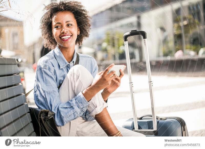 Happy young woman sitting on a bench with cell phone and suitcase human human being human beings humans person persons African black black ethnicity coloured 1