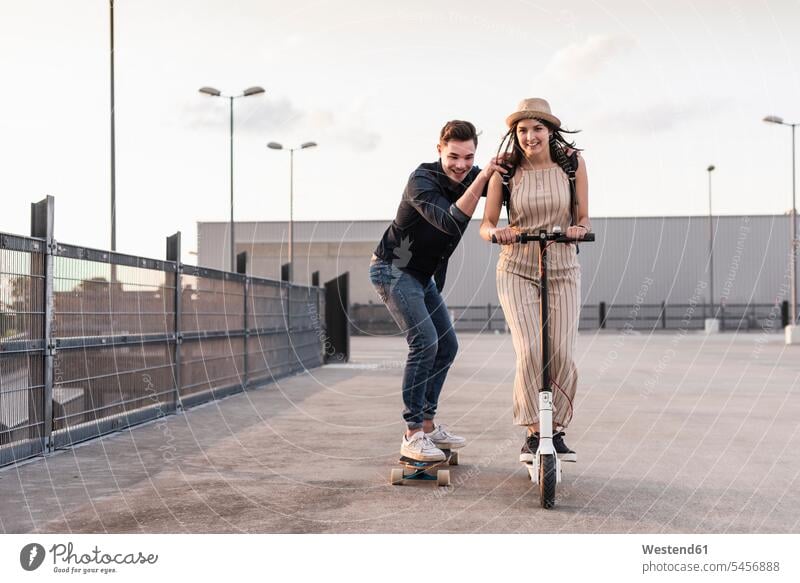 Young man and woman riding on longboard and electric scooter on parking deck human human being human beings humans person persons caucasian appearance