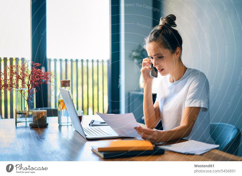 Businesswoman holding document talking over smart phone at desk in home office color image colour image casual clothing casual wear leisure wear casual clothes