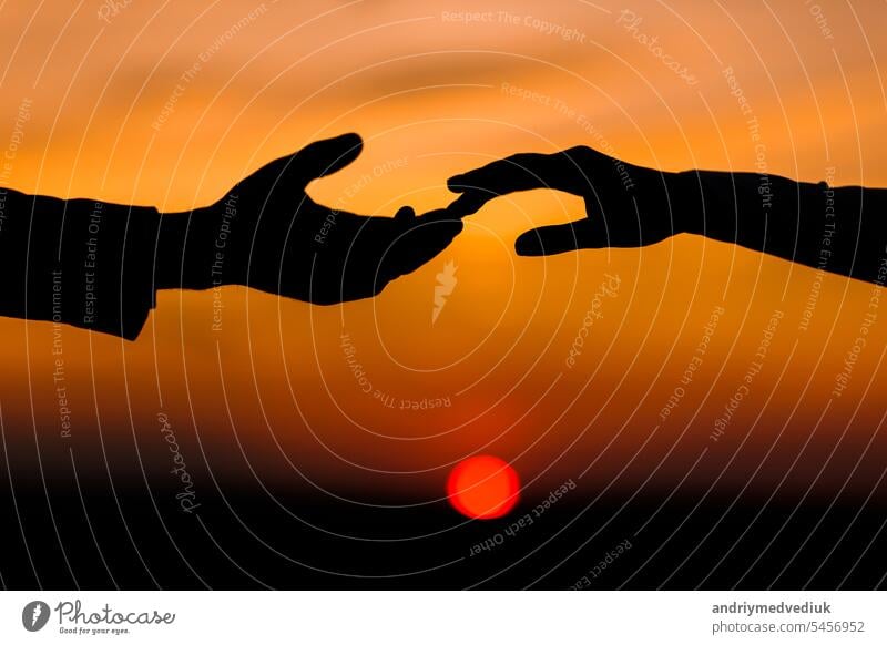 Valentine day. Silhouette of hands of woman and man reaching each other, touching fingers with tenderness on orange sunset sky background. Helping hands, save and support people concept. Friendship.