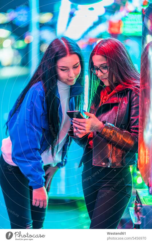 Two teenage girls sharing smartphone on a funfair at night friends mate female friend telecommunication phones telephone telephones cell phone cell phones