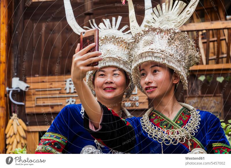China, Guizhou, two Miao women wearing traditional dresses and headdresses taking a selfie with smartphone Miao people Miao tribe Native Locals Formal wear