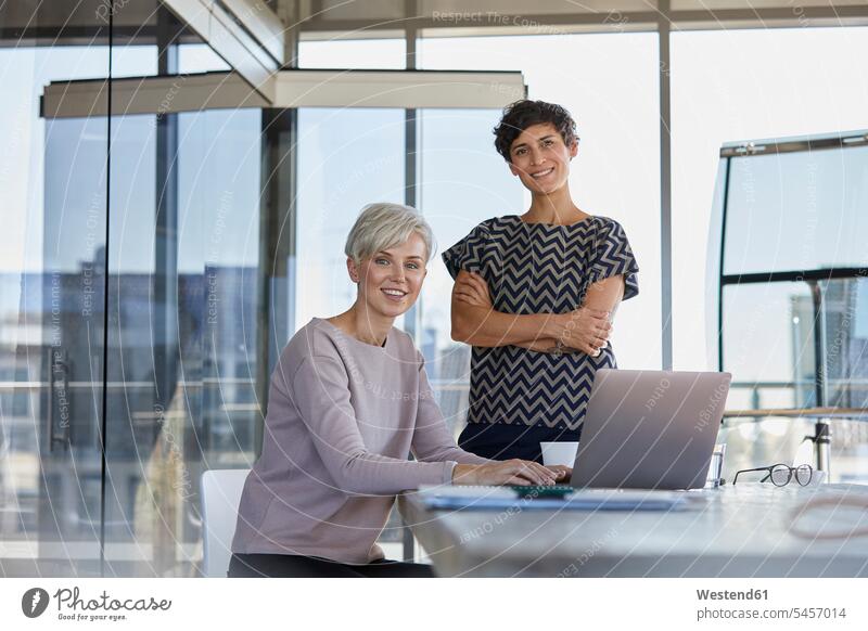 Portrait of two smiling businesswomen with laptop at desk in office desks businesswoman business woman business women offices office room office rooms