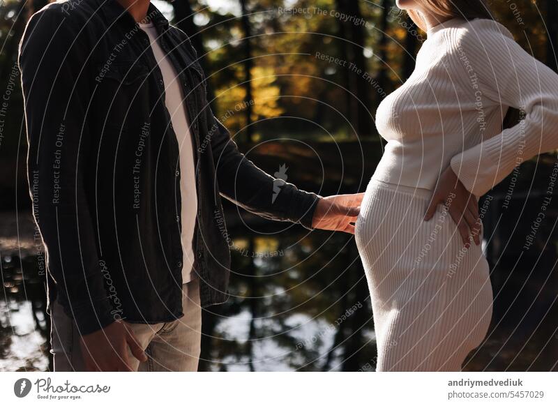 cropped photo of couple, man and pregnant wife are holding hands and walking by the lake on sunlight sunset. Happy family enjoying pregnancy on nature. The concept of Mother's Day and Women's Day.