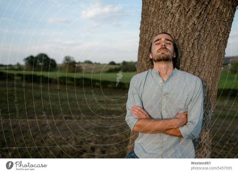 Relaxed young man leaning against a tree trunk in the countryside human human being human beings humans person persons caucasian appearance caucasian ethnicity