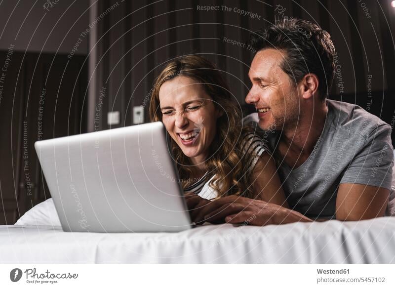 Happy couple lying on bed at home having fun with laptop Laptop Computers laptops notebook laying down lie lying down beds Fun funny twosomes partnership
