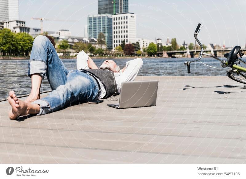Mature man with laptop and earbuds lying at the riverside in the city riverbank men males town cities towns earphones ear phone ear phones Laptop Computers