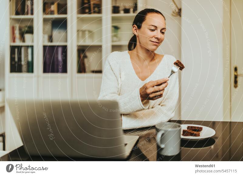Woman eating chocolate brownie while sitting with coffee and laptop in illuminated kitchen brownies Chocolate Chocolates Sweets Sweet Food Candies foods