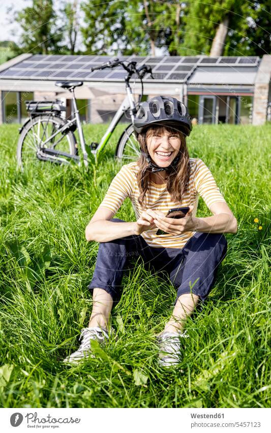 Laughing woman with bicycle using cell phone on a meadow in front of a house human human being human beings humans person persons caucasian appearance