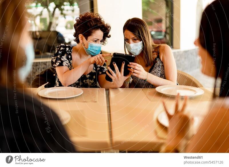 Women wearing masks while sharing digital tablet sitting with friends at table in restaurant color image colour image Spain indoors indoor shot indoor shots