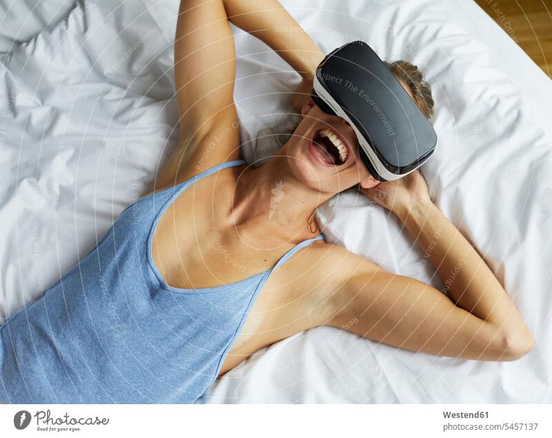 Laughing young woman lying in bed wearing VR glasses specs Eye Glasses spectacles Eyeglasses laughing Laughter females women beds happiness happy laying down