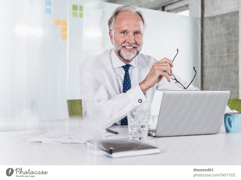 Portrait of smiling mature businessman sitting at desk in office with laptop desks Businessman Business man Businessmen Business men offices office room