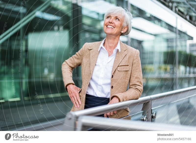 Smiling senior businesswoman leaning on railing in the city looking up businesswomen business woman business women rested on Railing Railings town cities towns